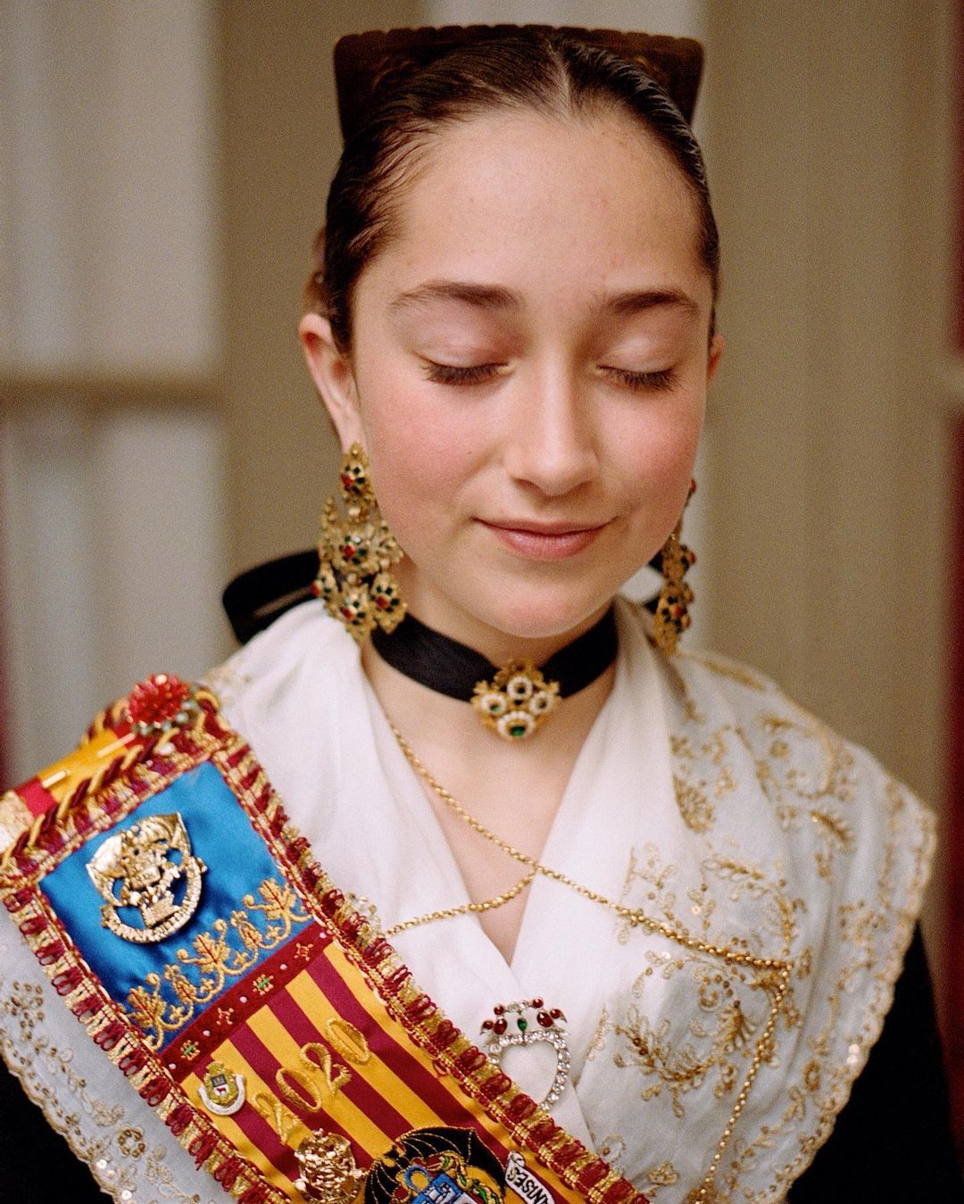 Close-up of a young woman with her eyes closed, wearing traditional attire with ornate jewelry and a colorful sash.
