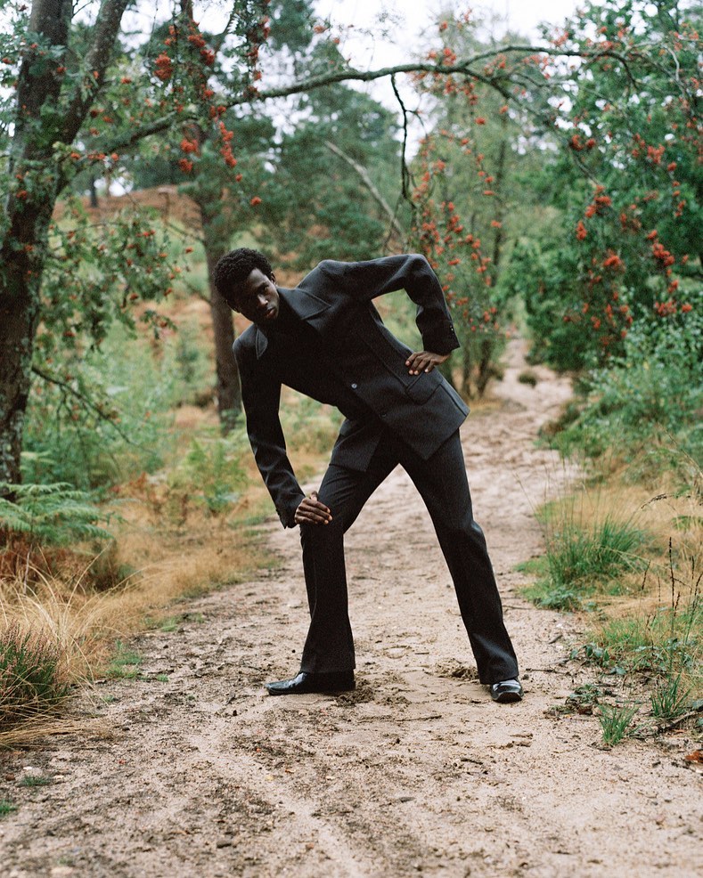 Adult male fashion model in a dark suit poses on a dirt path in a wooded area with green trees and red berries on branches overhead.