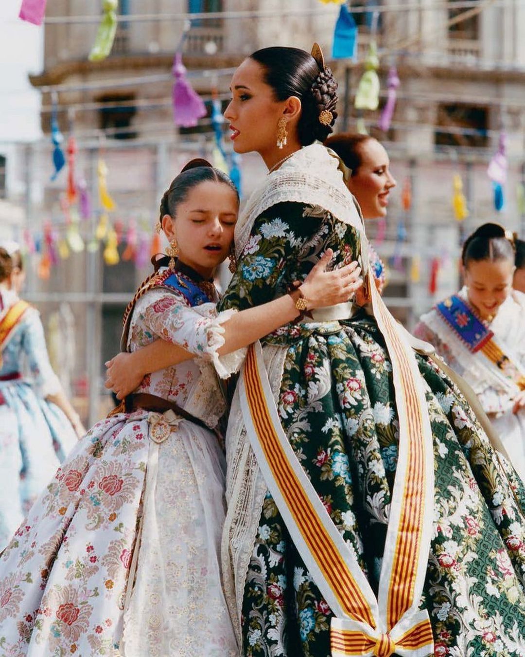 Two young girls in traditional Spanish dresses hug, celebrating Las Fallas festival in Valencia, Spain with colorful decorations.