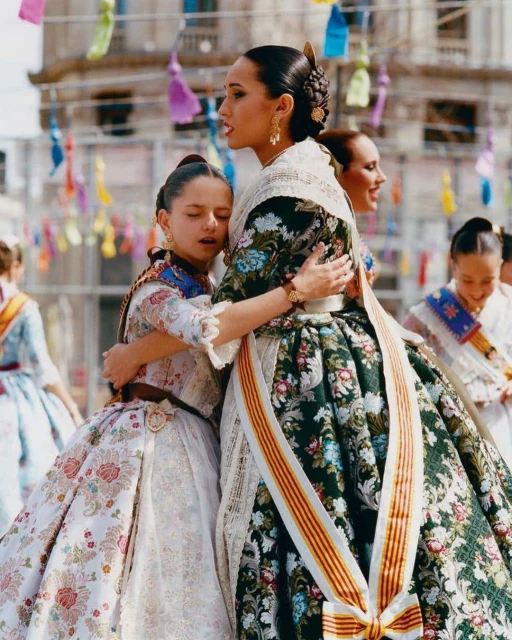 Two young girls in traditional Spanish dresses hug, celebrating Las Fallas festival in Valencia, Spain with colorful decorations.