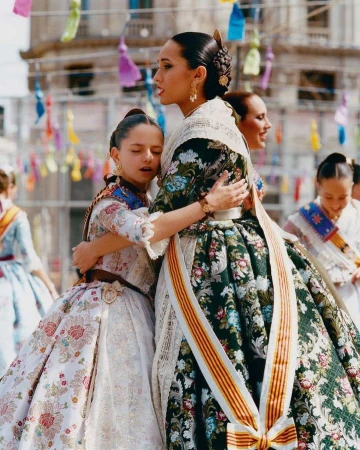 Two young girls in traditional Spanish dresses hug, celebrating Las Fallas festival in Valencia, Spain with colorful decorations.