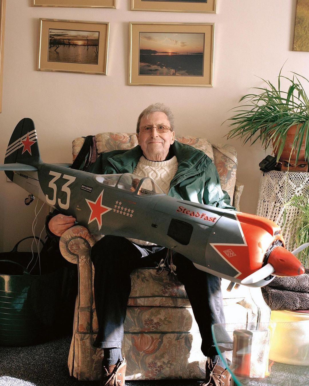 Elderly man sits indoors in an armchair holding a large model airplane with red and grey markings. Framed art hangs on the wall behind him.