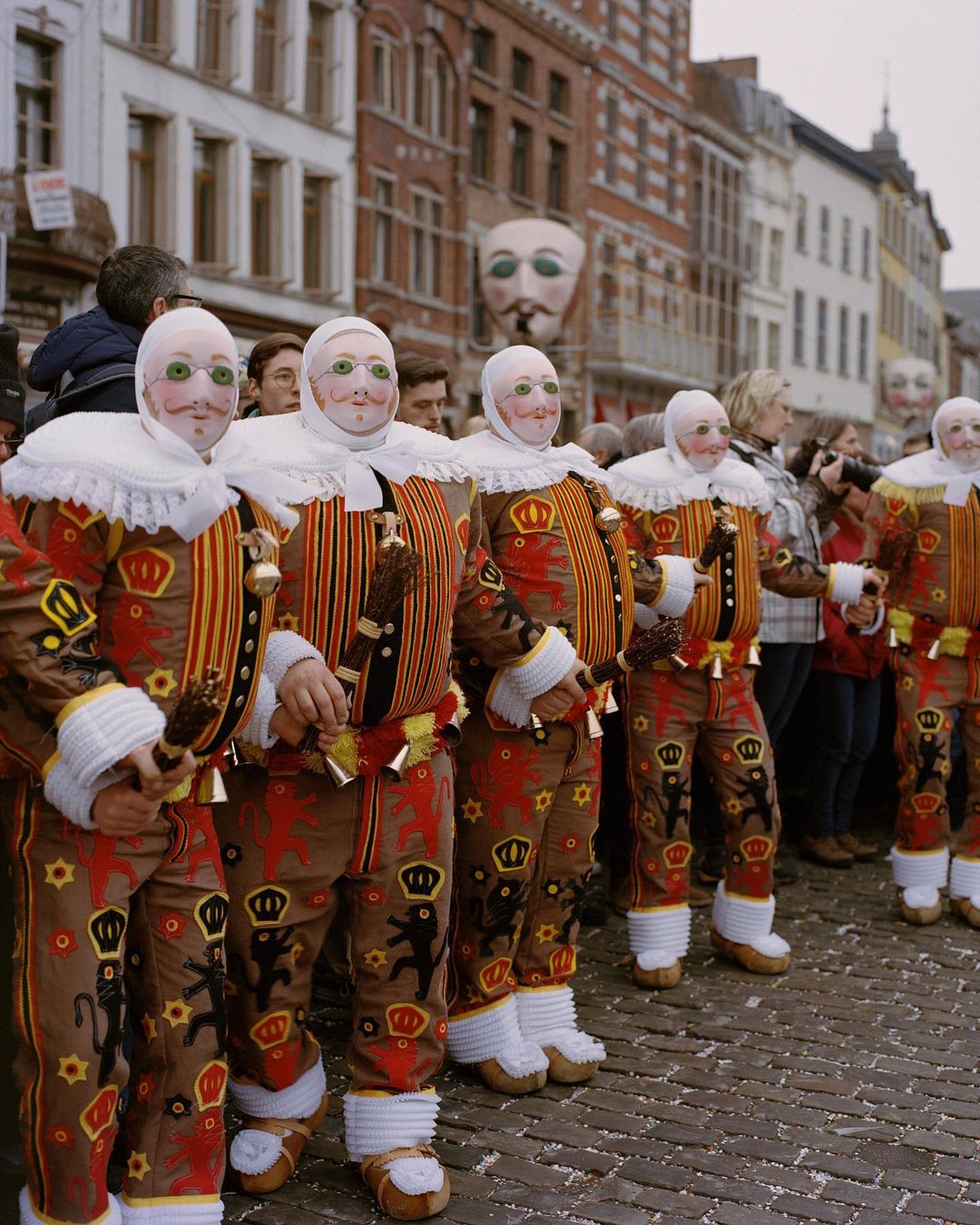 Belgian carnival performers in patterned costumes and masks stand in a line on a brick street in a European urban setting.