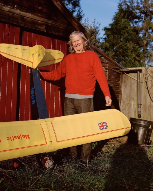 Adult man poses outdoors next to a vintage yellow model airplane near a red barn, with trees and a blue sky in the background.