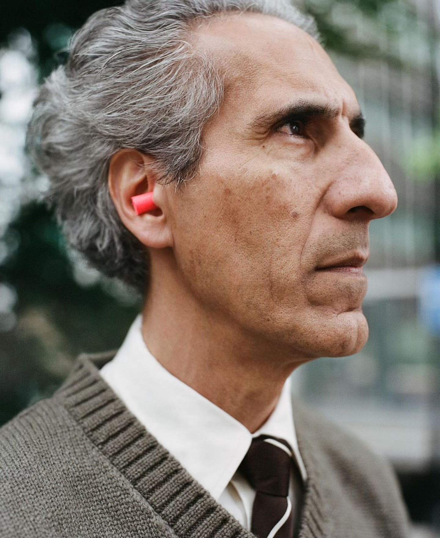 Profile of adult male with earplugs outdoors. He has gray hair and wears a sweater, shirt, and tie. Building and foliage visible in background.