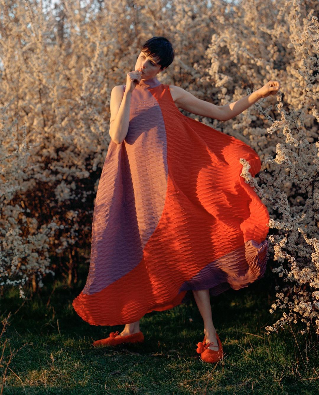 A woman with short dark hair stands outside wearing a pink and orange dress with matching flats against a flowering bush.