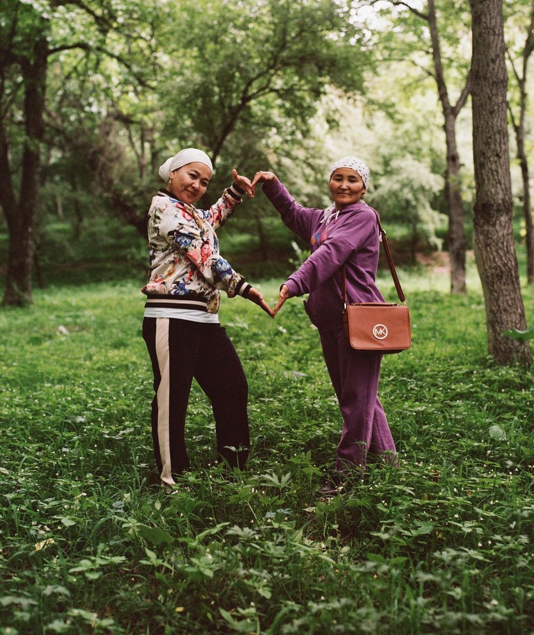 Two women standing in a lush green park making a heart shape with their hands, photographed by Hill & Aubrey.
