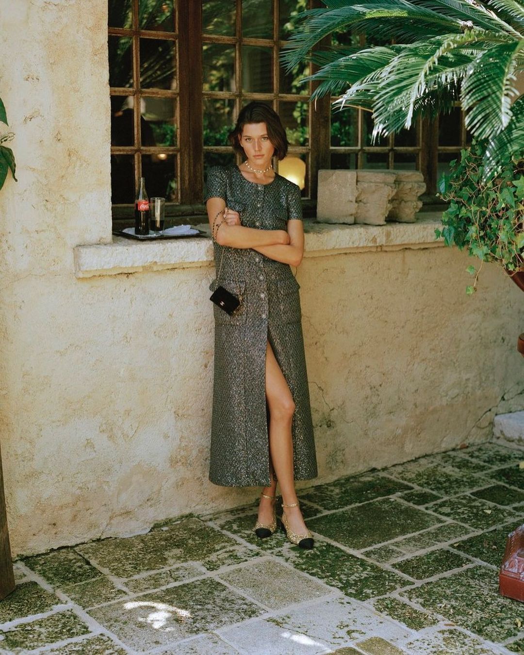 A woman poses outdoors in a tweed button-down dress with a purse. Sunlight and shadows create a natural, textured scene in neutral colors.