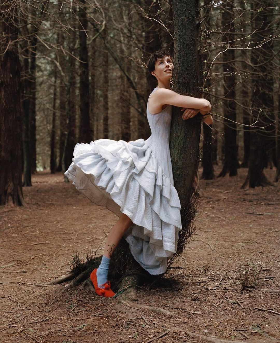 A woman in a light blue dress stands hugging a tree in a forest, with orange shoes and blue socks adding a pop of color to the scene.