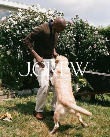 Adult male interacts with a golden Labrador Retriever in a grassy yard with blossoming bushes and trees on a sunny day.