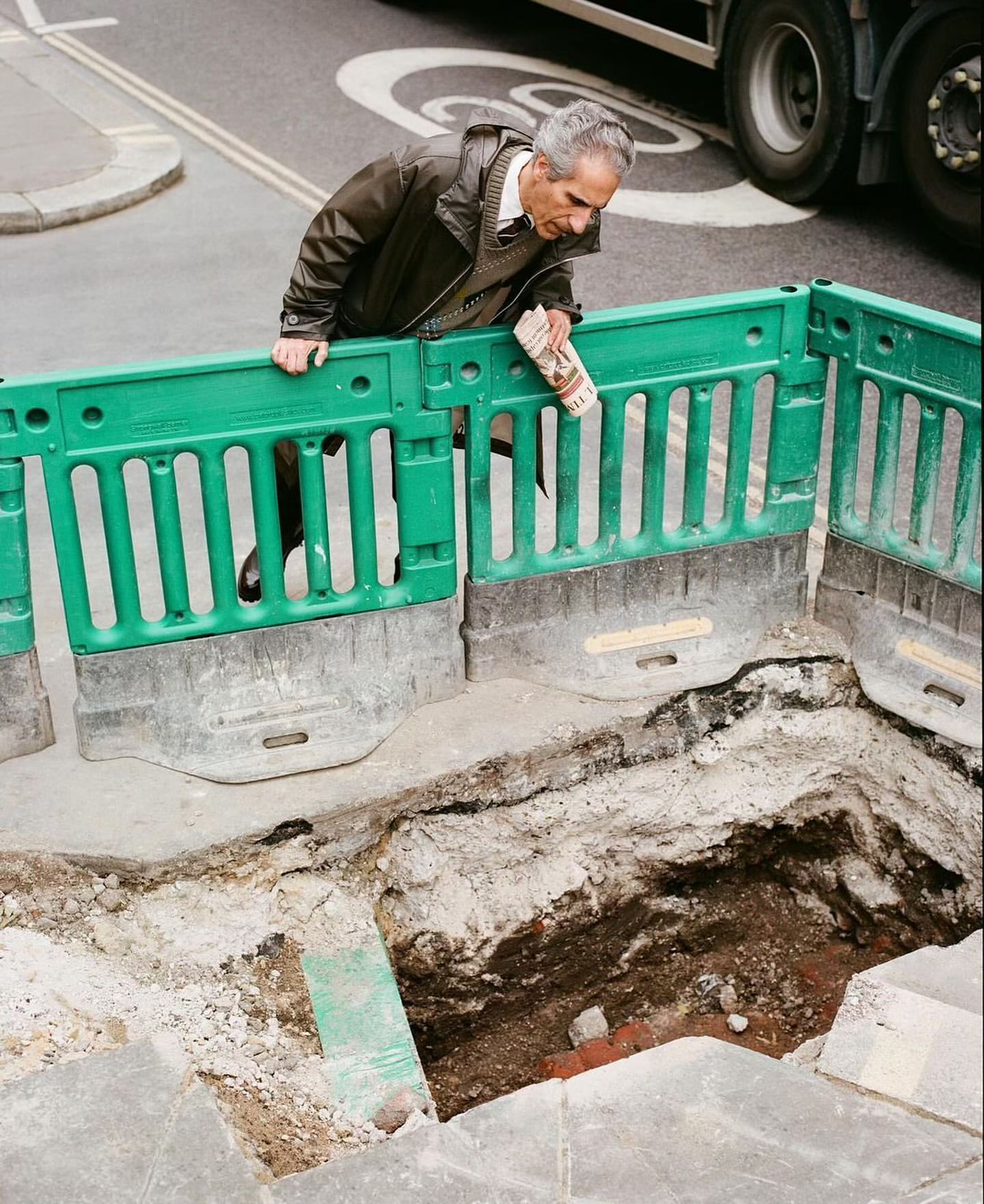 Man reads newspaper near open construction pit on city street with truck traffic. Safety fence and concrete barrier at edge of hole.