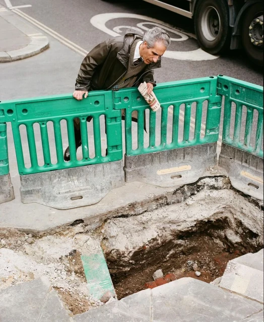 Man reads newspaper near open construction pit on city street with truck traffic. Safety fence and concrete barrier at edge of hole.