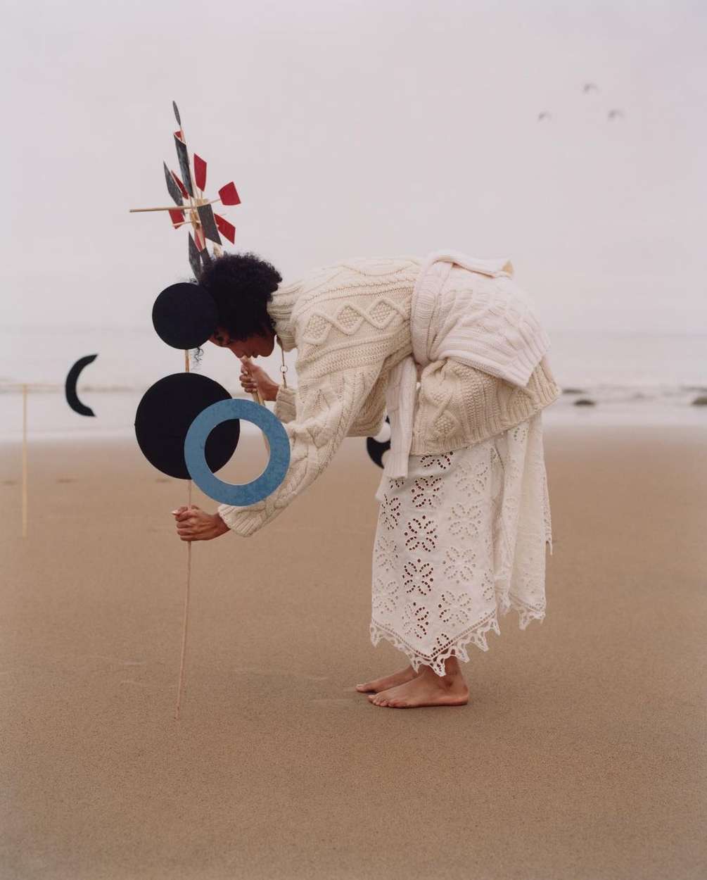 A woman in a white sweater and lace skirt is on a sandy beach, appearing to look at or interact with an abstract sculpture.