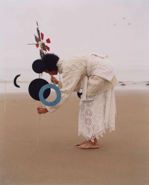 A woman in a white sweater and lace skirt is on a sandy beach, appearing to look at or interact with an abstract sculpture.