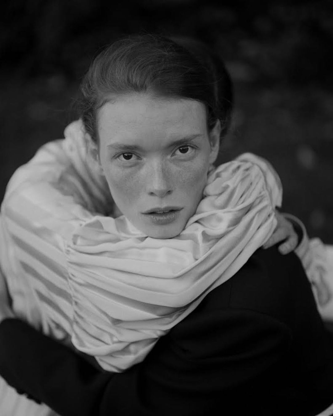 Black and white portrait of a woman embracing a person from behind in an outdoor setting with diffused, soft light.