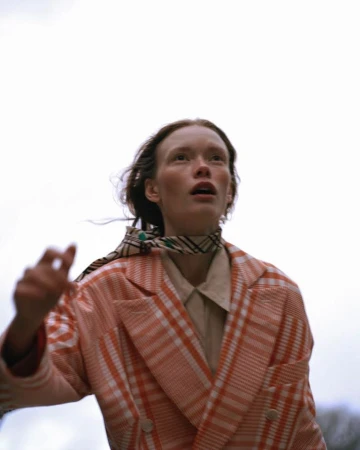 Model in an orange and white plaid double-breasted coat and patterned neck scarf looking upward outdoors in New York.
