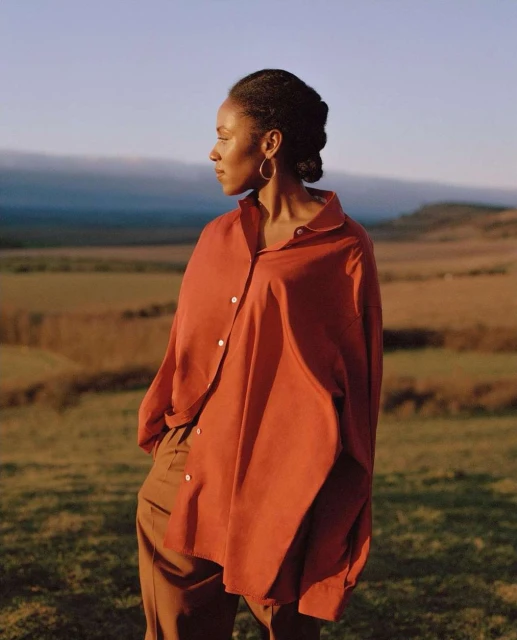 A woman with an orange button-down shirt stands outdoors in a field at sunset, looking off to the side with a serene expression.