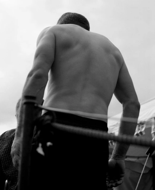 Black and white shot of a muscular man standing in what appears to be a boxing ring, facing away from the camera. 