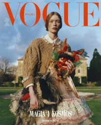 Model holding a colorful horned mask wearing a textured camel cardigan in an outdoor garden setting with a large building.