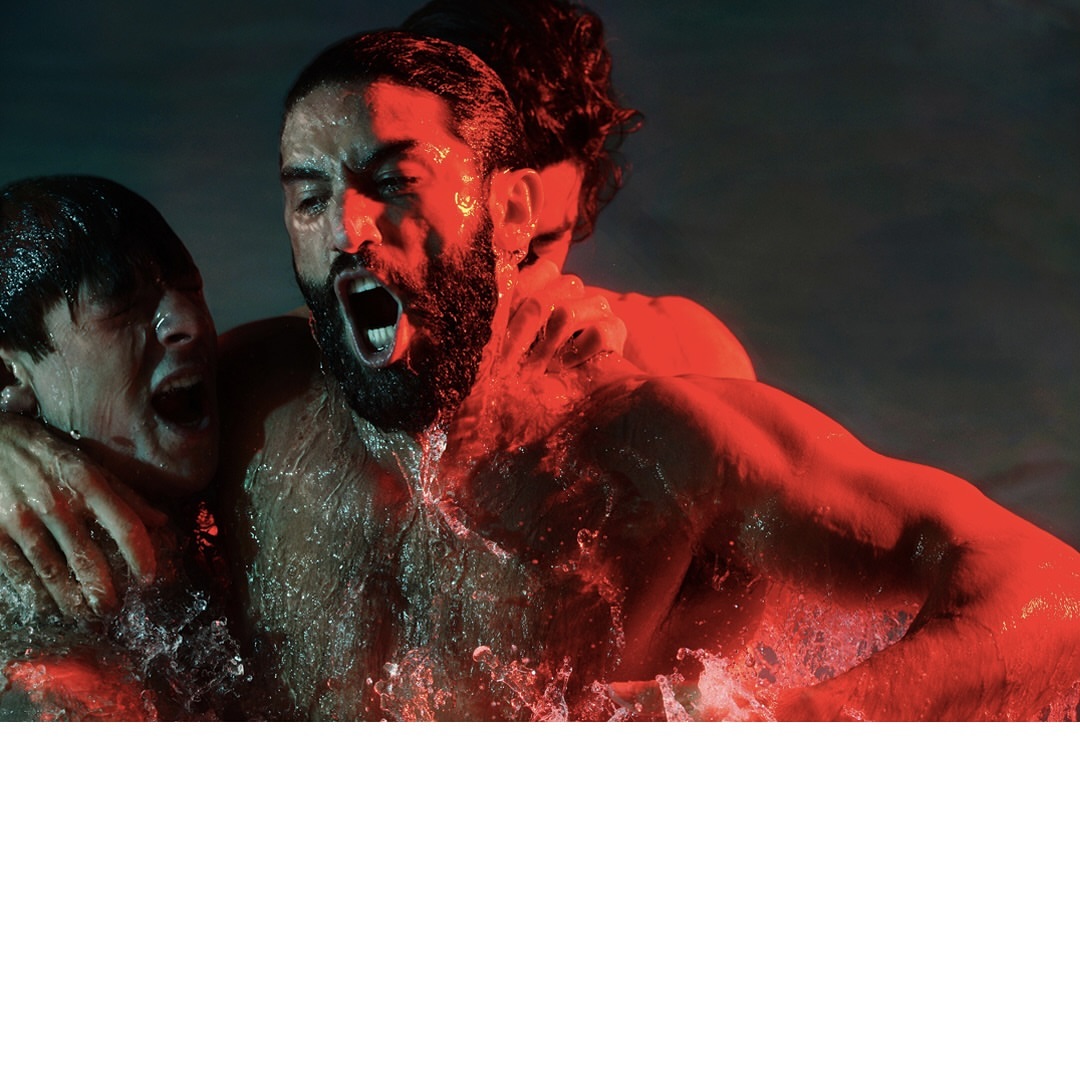 Three adult men in water, faces expressing anguish, water splashing, red and green lighting creating a dramatic scene.
