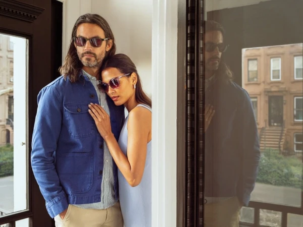 Couple models stylish eyewear outdoors. Man in blue jacket, woman in light dress, both wear sunglasses in an urban setting with brick buildings.