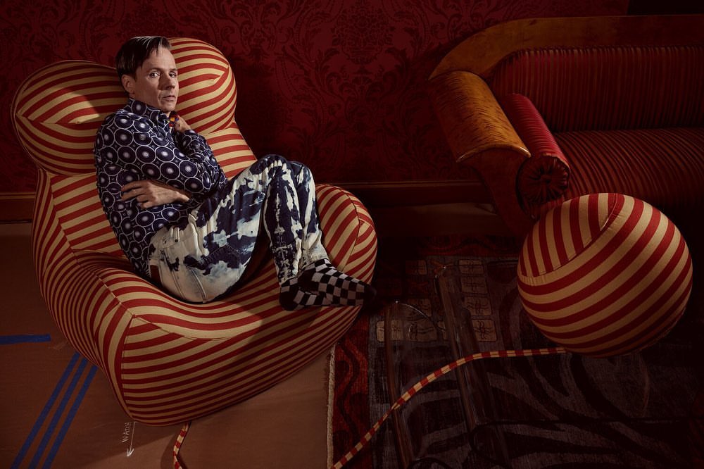 Man seated in striped armchair with patterned shirt and pants, set against a decorative red wall and ornate carpet in a photo studio.