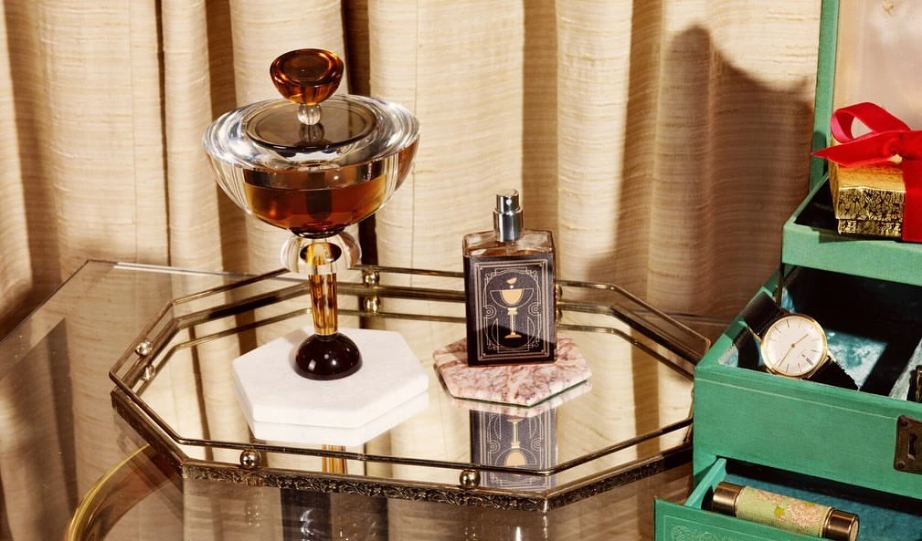 Still life of a glass decanter with amber liquid, a perfume bottle, and a green jewelry box with a red ribbon on a mirrored tray.