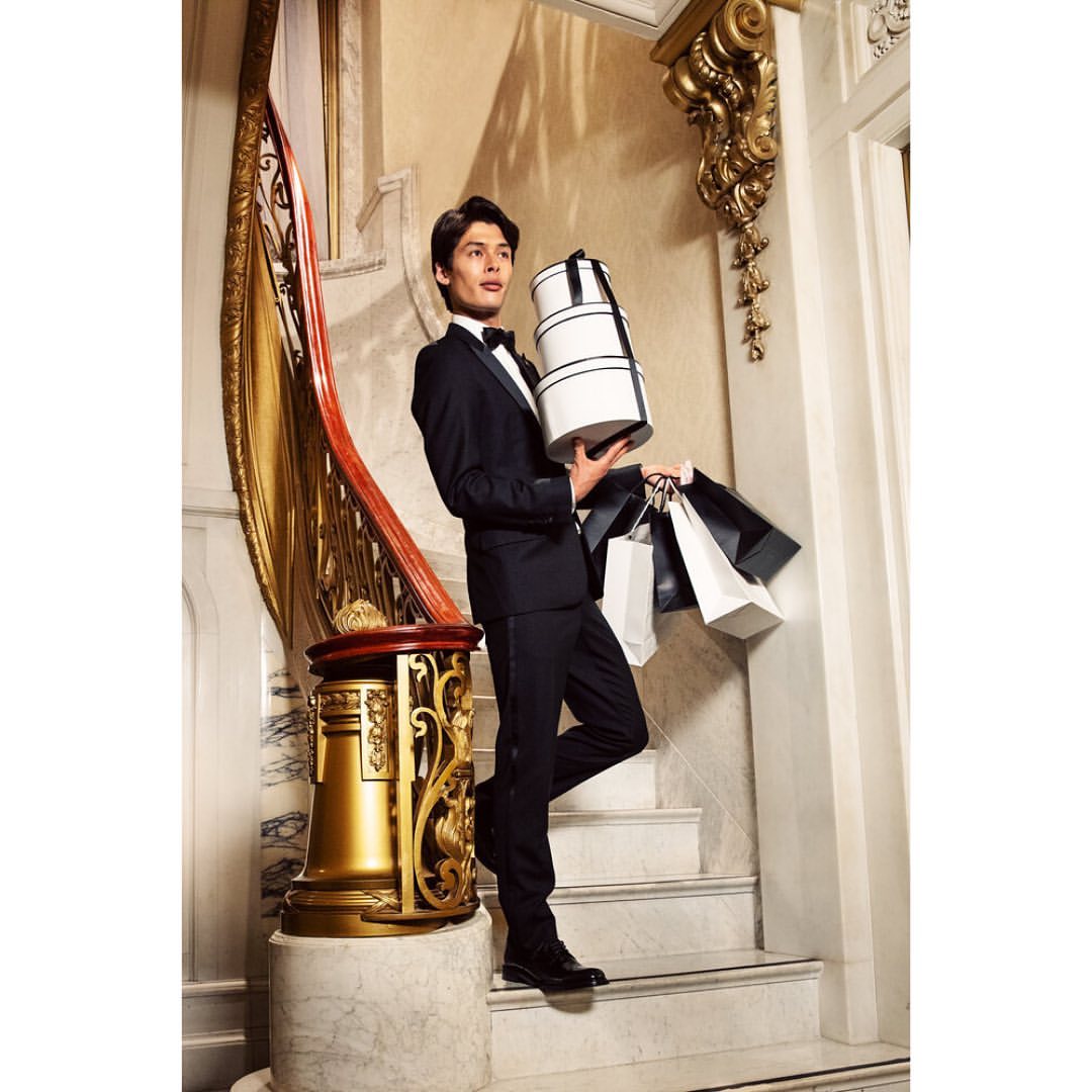 Elegant man in tuxedo on marble staircase holding shopping bags and boxes with decorative gold gilded railing and neutral walls.