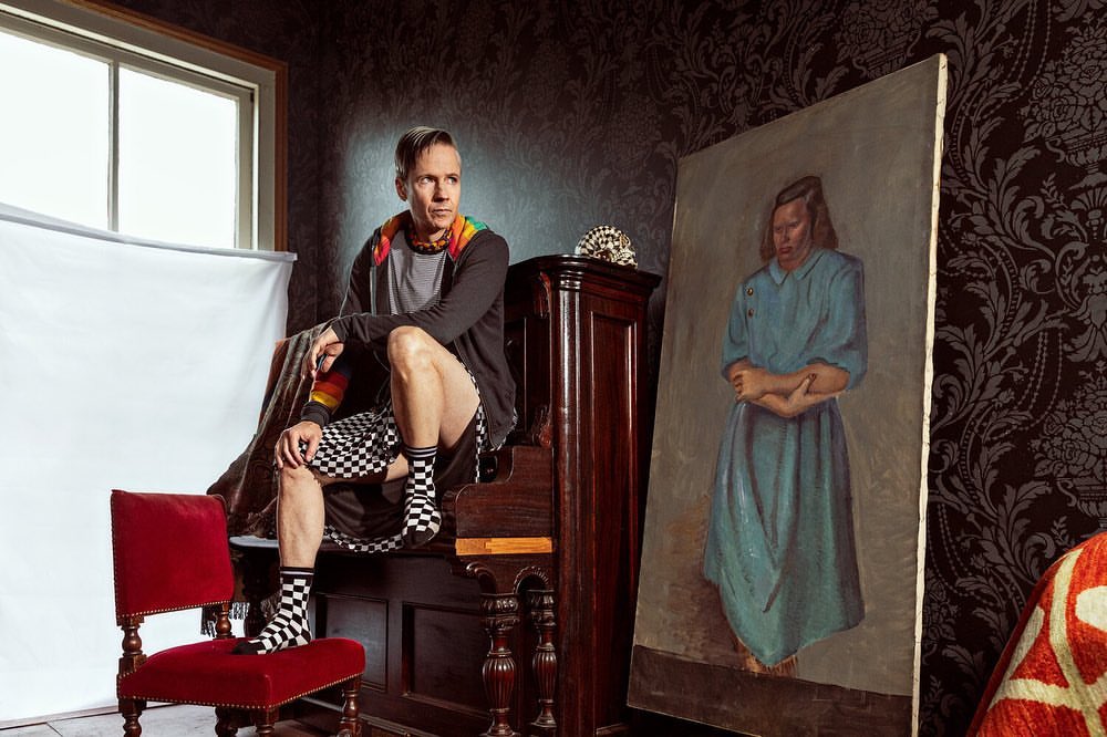 Adult male seated on a piano, wearing a gray hooded sweatshirt and checkered shorts and socks, next to a framed painting in an indoor setting.