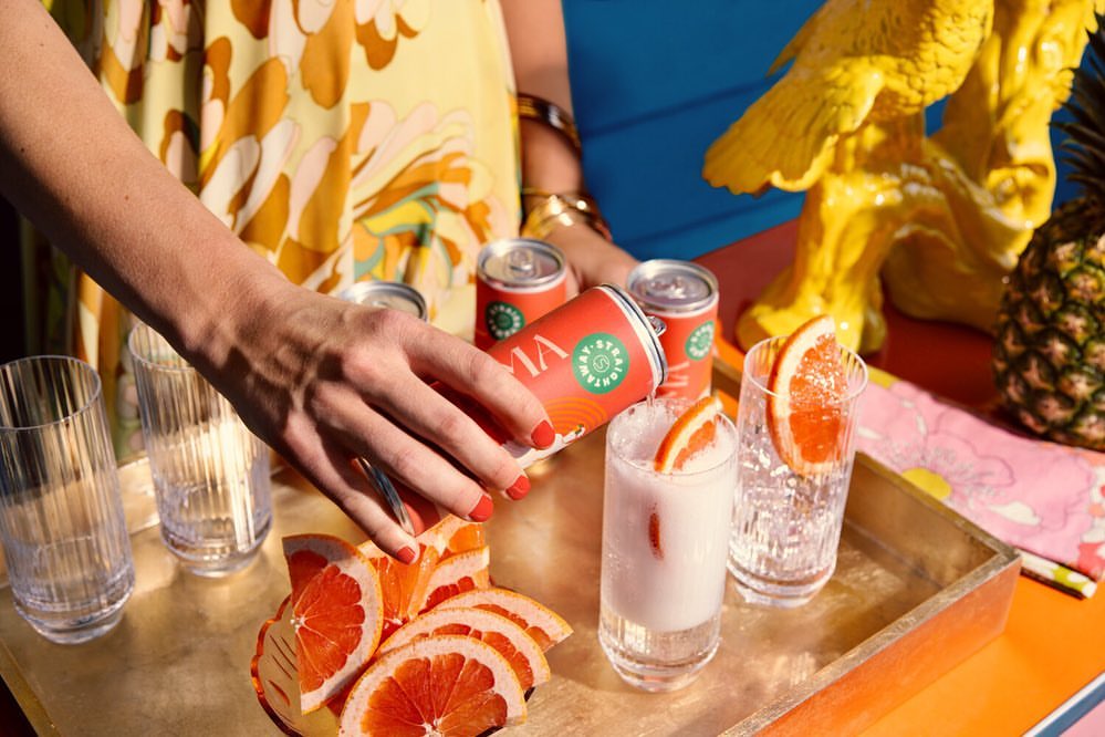 A person pours a Viva sparkling beverage into a glass with grapefruit slices, featuring vibrant yellows and oranges in a cheerful setting.