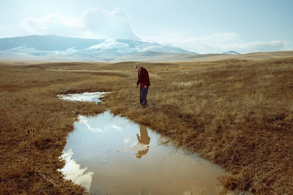 An older adult male stands near a water stream in a golden field, his reflection mirroring in the water under a bright sky and distant mountains.