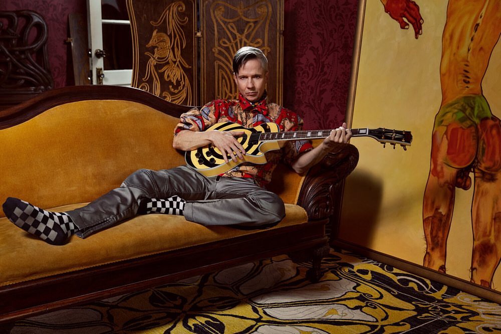 Man with silver hair poses with an electric guitar on an antique gold couch in front of a nude art painting and patterned wall.