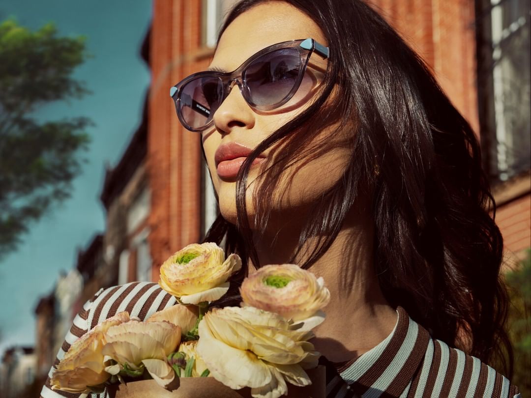 Brunette woman wearing sunglasses holds ranunculus flowers against a brick building and blue sky backdrop in an eye-level outdoor shot.