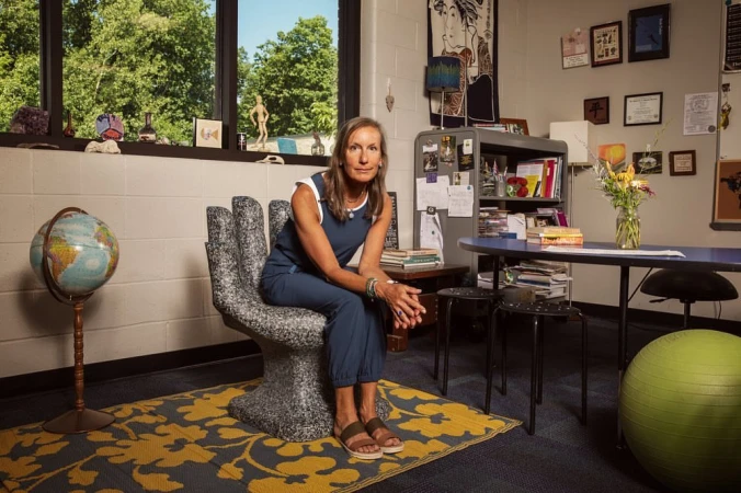 Woman in blue jumpsuit sitting on a hand-shaped chair in a classroom with a globe, desk, and bright sunlight streaming through the window.