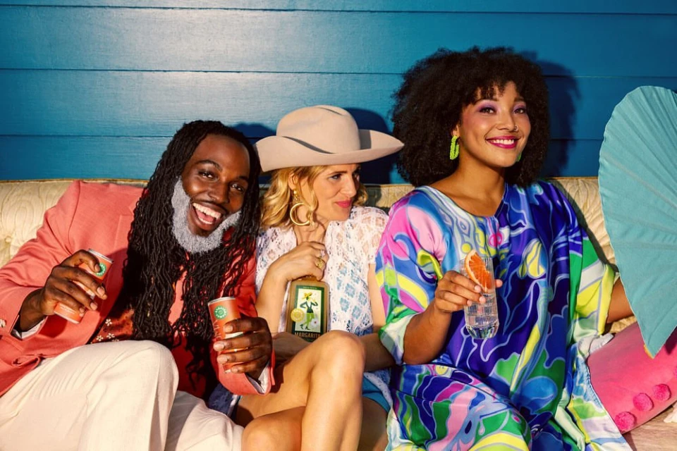 Three smiling people—two women and a man—sit indoors on a couch holding canned beverages and a bottle. They wear colorful clothes.