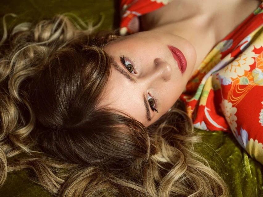 Close-up portrait of a fair-skinned adult woman with long, wavy, blonde and brown hair lying on a green velvet surface, wearing a red floral robe.