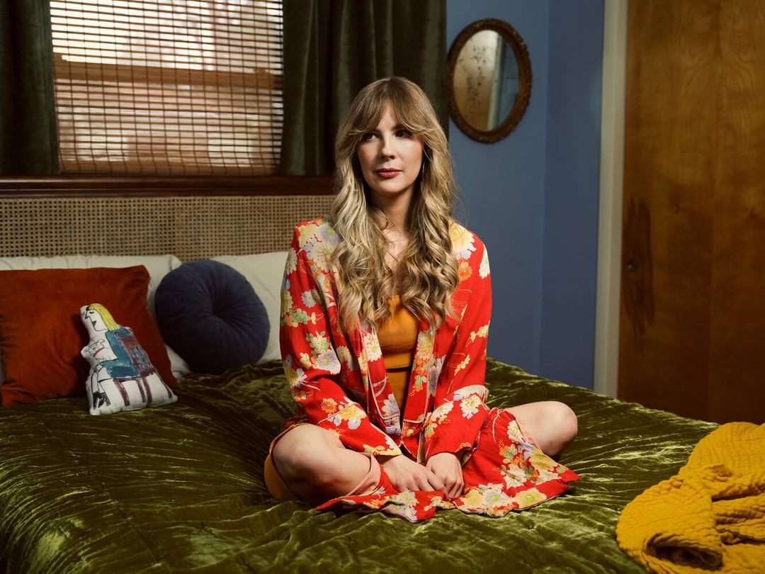Woman in floral robe sits cross-legged on a green velvet bed with decorative pillows, lit by soft, natural light from a nearby window.