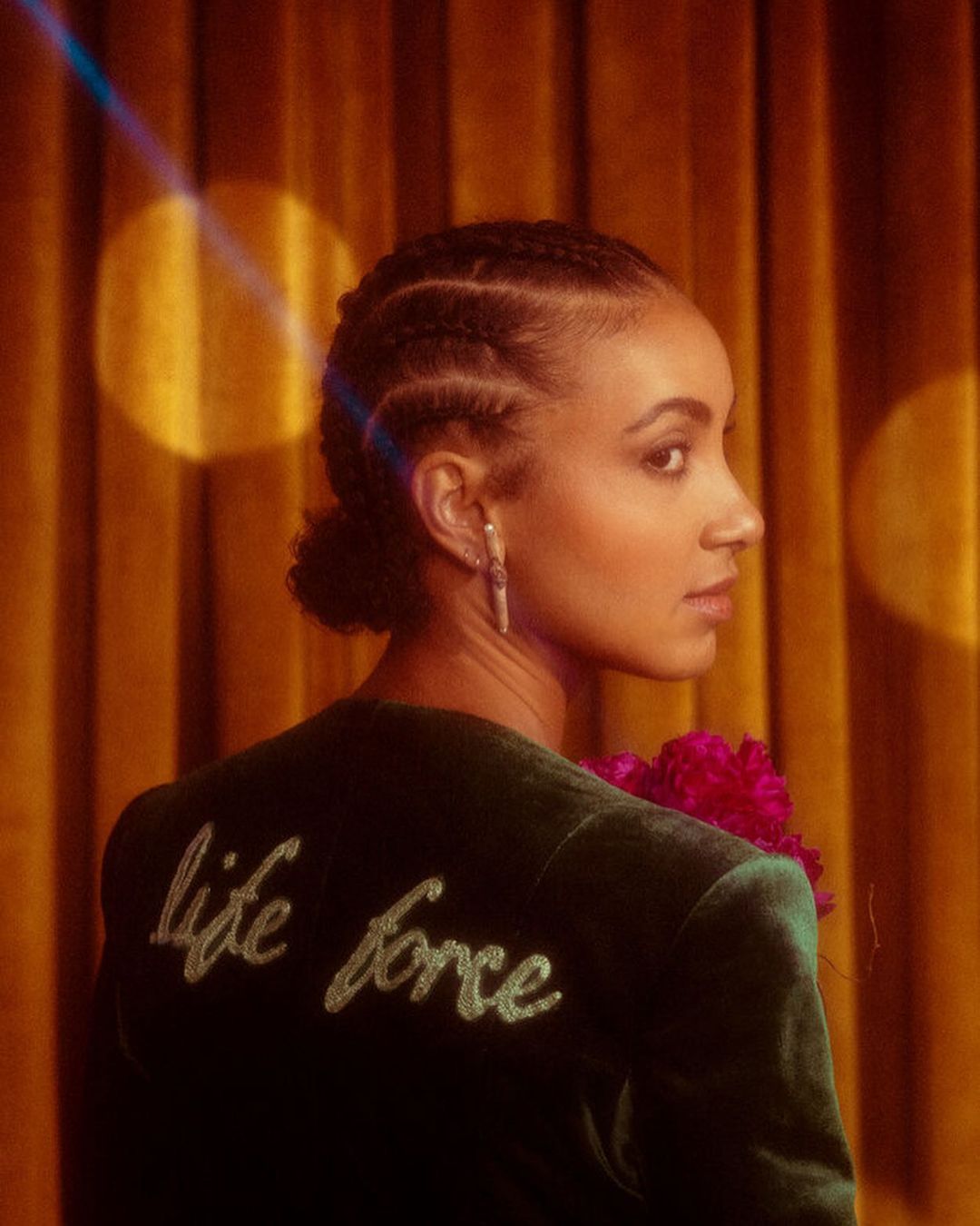 Elegant portrait of an adult woman wearing a velvet blazer with 'life force' embroidered on the back, standing in front of a curtain.