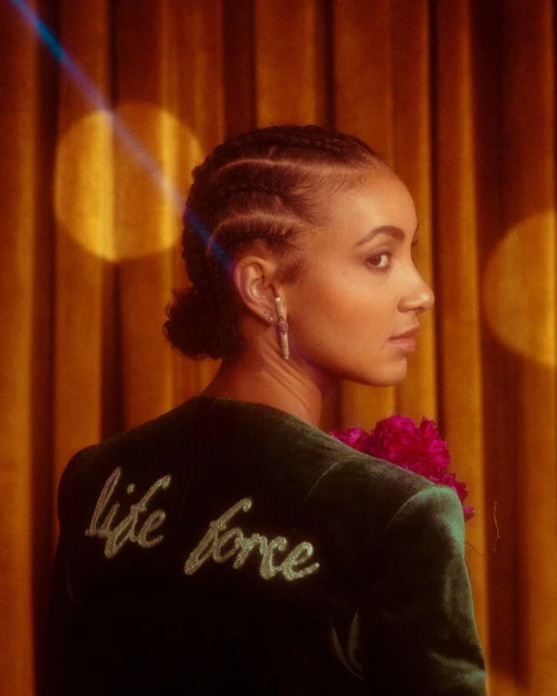 Elegant portrait of an adult woman wearing a velvet blazer with 'life force' embroidered on the back, standing in front of a curtain.