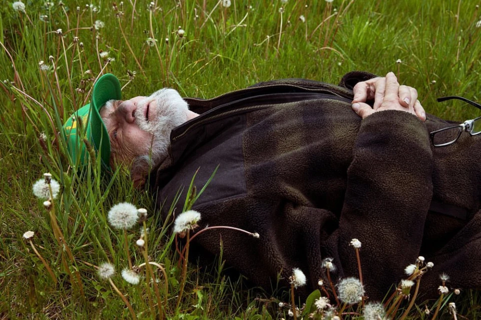 Elderly man with white beard and green hat relaxes lying in tall green grass with dandelions, wearing a brown fleece jacket outdoors.