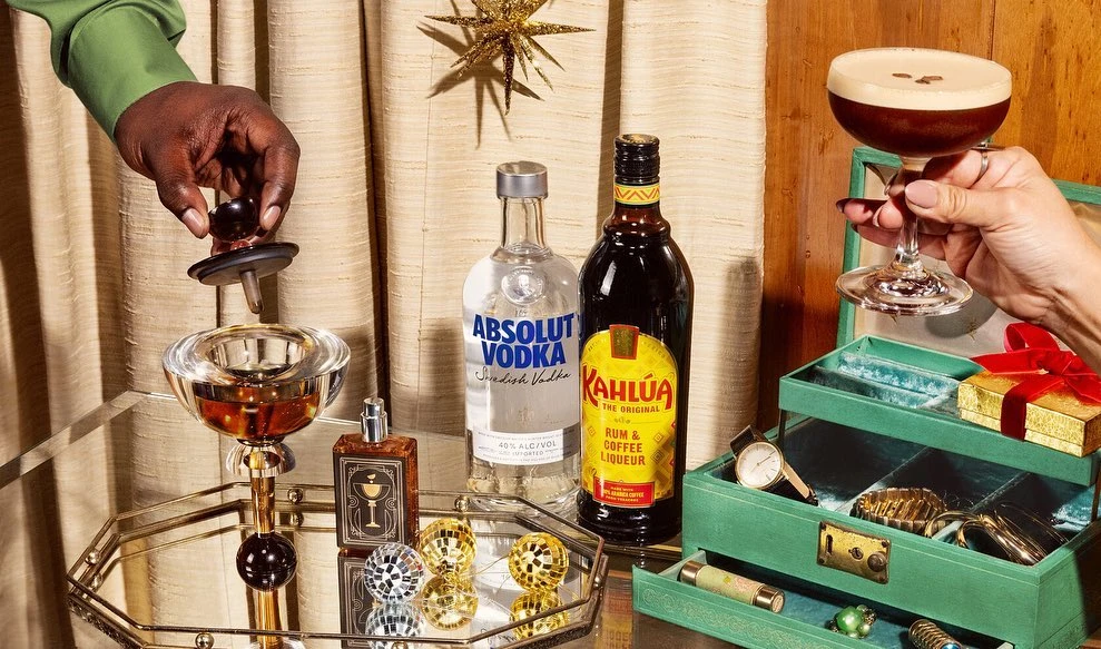 Cocktail preparation scene with hands, liquors, and jewelry box on mirrored tray against neutral draped fabric and golden star.