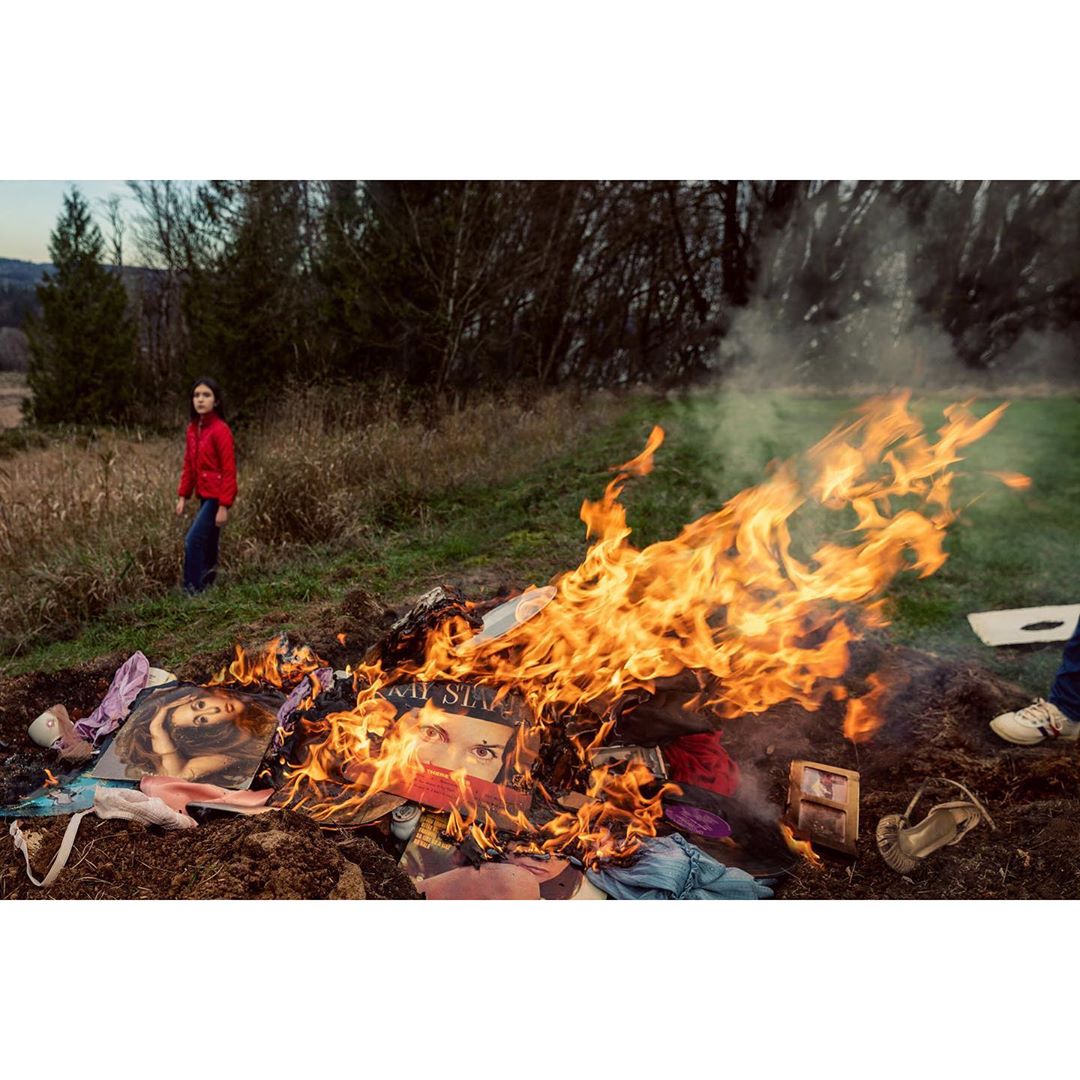 Person stands near a fire consuming printed materials outdoors with grass and trees in the background on a sunny day.