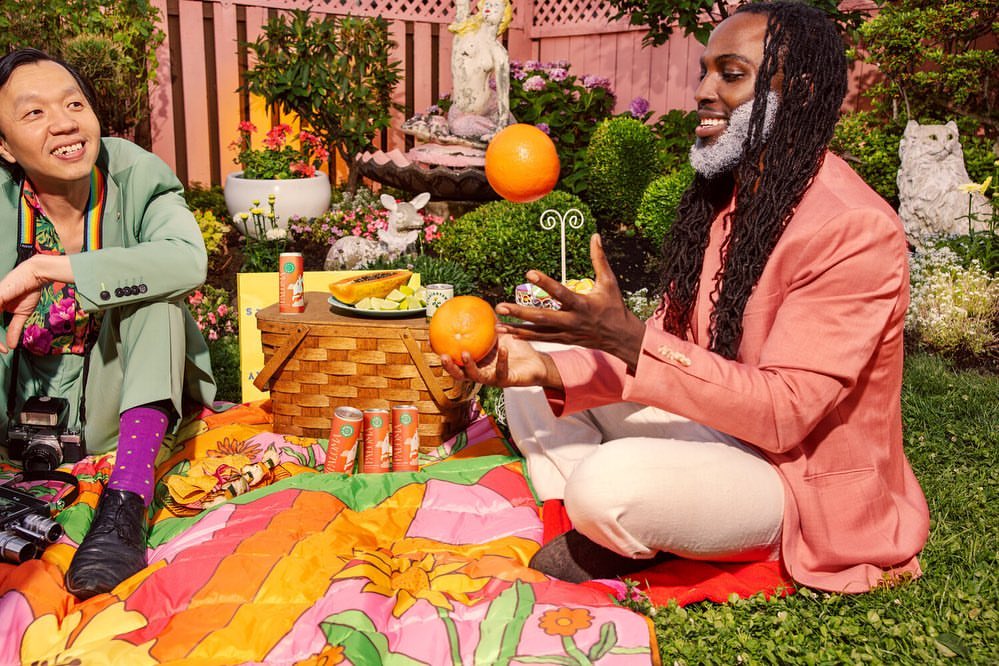 Two men at an outdoor picnic with fruit, cans, and vintage cameras, sitting on a colorful blanket on the grass. Lush greenery.