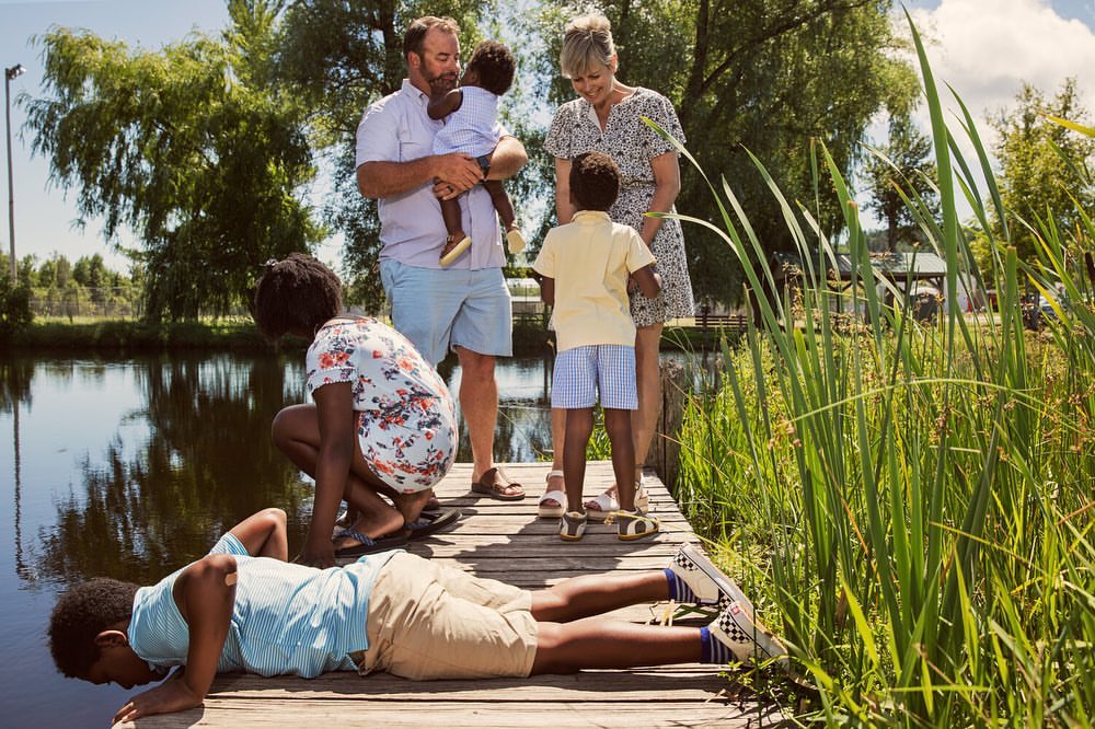 A diverse family stands and kneels on a wooden dock near a pond on a sunny day; a man holds a toddler; trees and tall grasses surround them.