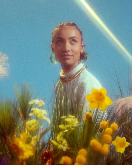 Portrait of a young woman with braided hair and gold jewelry in a field of yellow daffodils against a blue sky with lens flare.