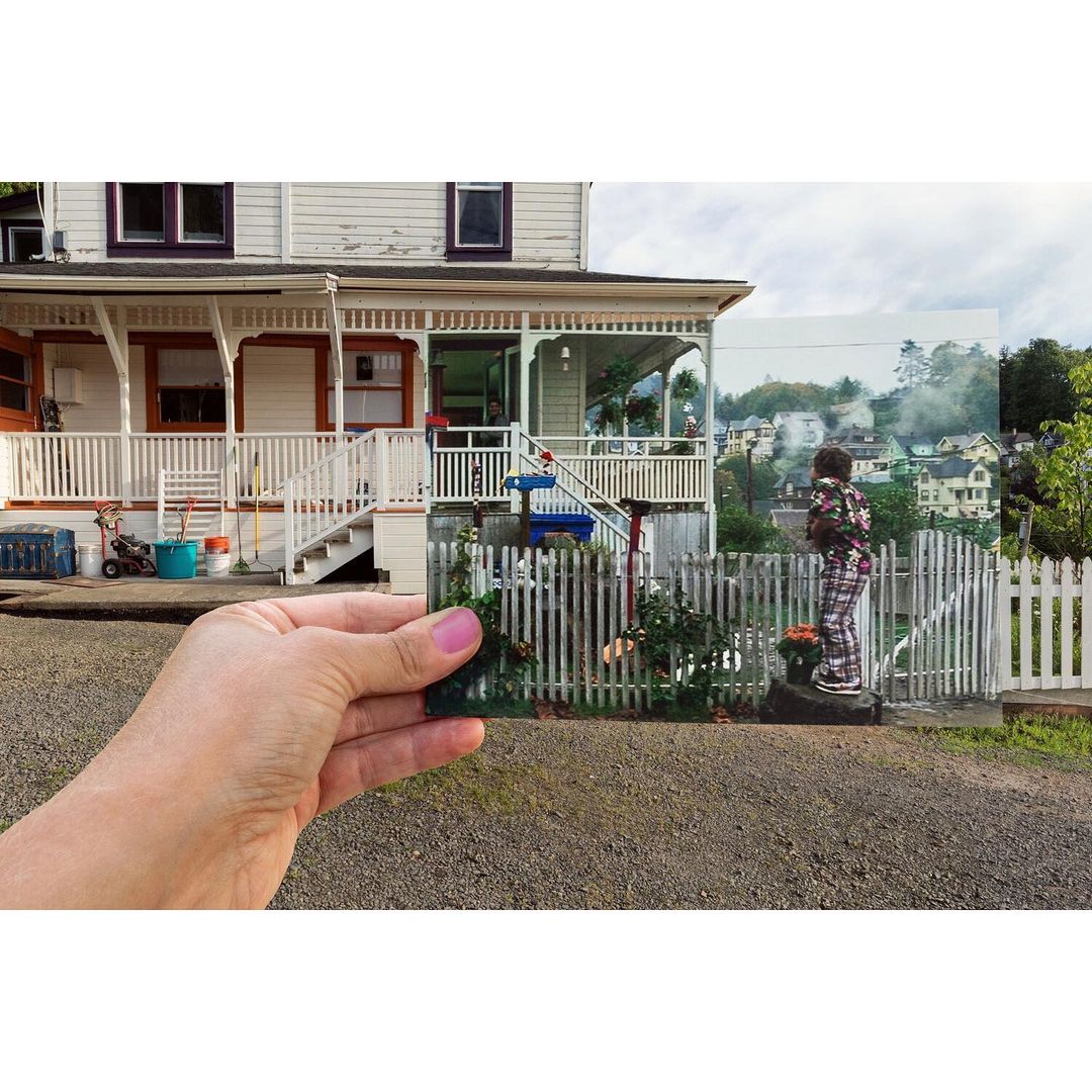 Youngster gazes at a quaint neighborhood captured in a scenic card held by a hand in front of a white house and gravel drive.