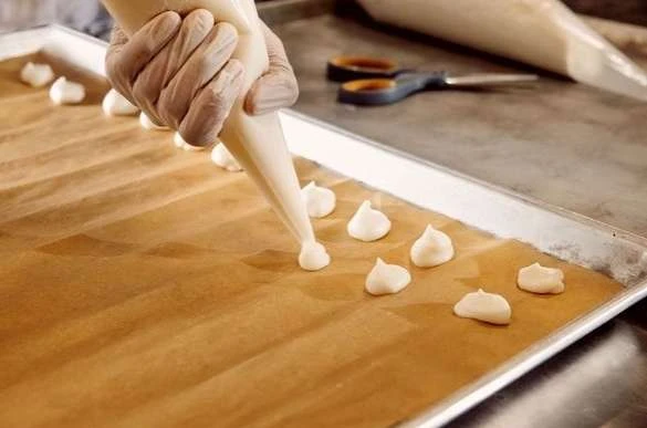 Hand piping meringue kisses onto parchment paper on a baking sheet with scissors and additional pastry bag in background.