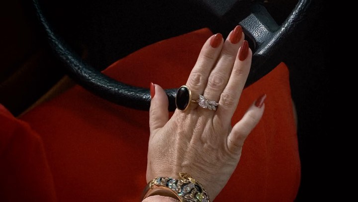Close-up shot of an adult woman's hand wearing red nail polish, multiple rings, and a bracelet while holding a black steering wheel.