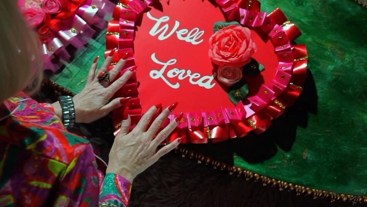 A woman with red nails and colorful top arranges a handmade red heart sign decorated with a rose and ribbon, reading, 'Well Loved'.