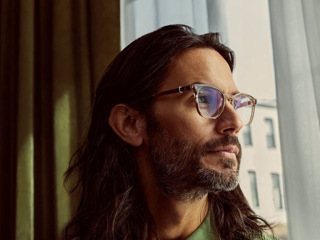 Man with long hair and beard wearing glasses looking out window. Close-up portrait of a man with green collared shirt inside.